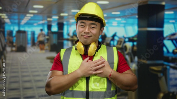 Fototapeta Man in gym wearing hard hat and reflective vest with headphones smiling and crossing hands on chest, expressing gratitude amid exercise equipment in bright indoor setting.