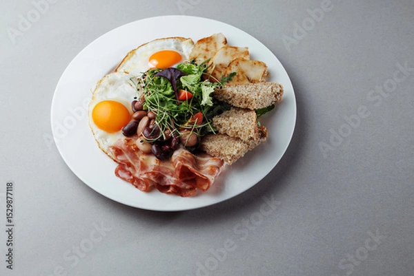 Fototapeta Healthy breakfast with eggs, bacon, grilled chicken, beans, salad and bread on white plate – top view gourmet morning meal on gray background