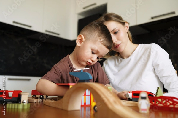 Fototapeta Young mother and her little son deeply focused while playing with a wooden toy train set at the kitchen table, sharing a quiet and thoughtful moment together.