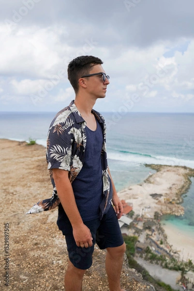 Obraz A young man in sunglasses and a tropical shirt stands on a cliff overlooking the ocean and Melasti beach on a cloudy day in Bali, Indonesia