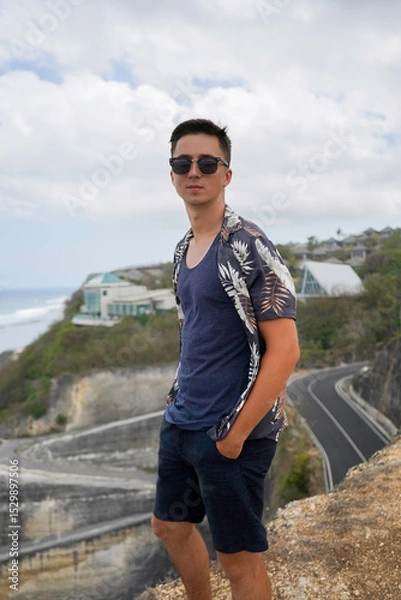 Obraz A young man in sunglasses and a tropical shirt stands on a cliff overlooking the ocean and Melasti beach on a cloudy day in Bali, Indonesia