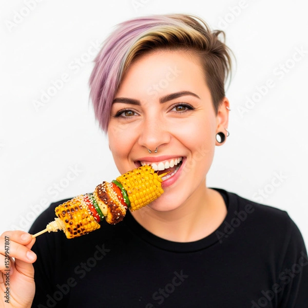 Fototapeta A smiling vegan woman with playful, short, colorful hair and a nose piercing joyfully eats a succulent skewer with a roasted corn cob against a stark white background.