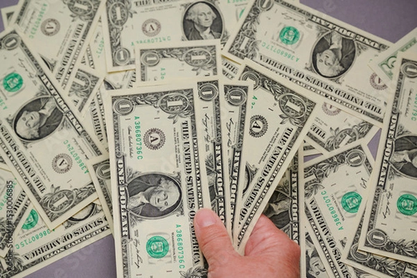 Fototapeta A hand holds several one dollar bills while others are scattered on a flat surface.