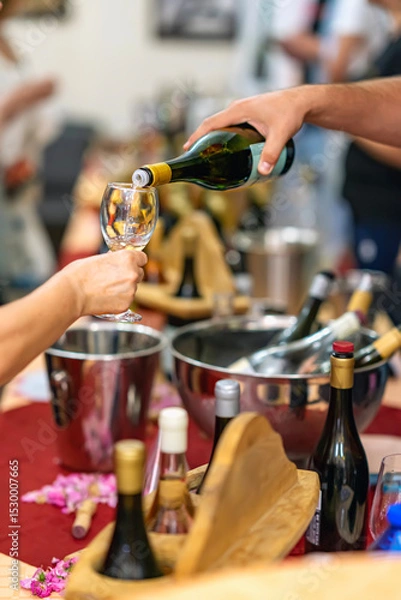 Fototapeta Wine tasting event with hand pouring white wine into glass, surrounded by wine bottles, ice buckets and wooden trays. Concept of celebration, sommelier service and premium lifestyle