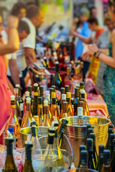 Fototapeta Wine Festival. People gathered at busy wine tasting festival table filled with various bottles and ice buckets. Concept of celebration, public event, wine party