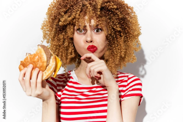 Fototapeta Young woman with curly hair in stripes holding a burger, playfully shushing with a surprised expression, ideal for healthy eating and fast food concepts