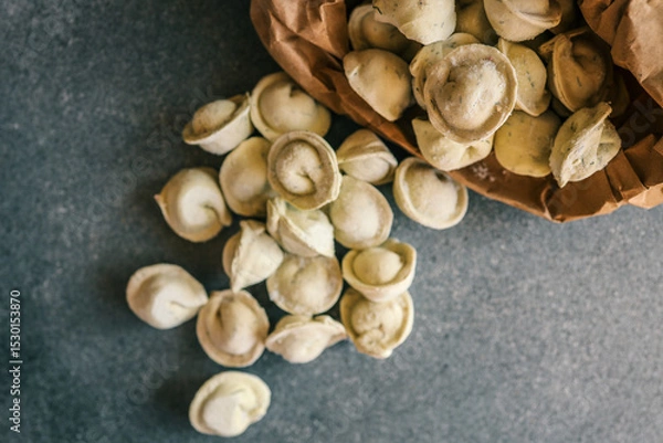 Obraz Frozen tortellini pasta spilling out of a brown paper bag onto a dark stone surface, ready for cooking or packaging