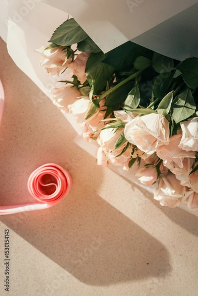 Obraz Elegant bouquet of pale pink roses wrapped in white paper with a pink ribbon roll nearby on light textured surface in sunlight