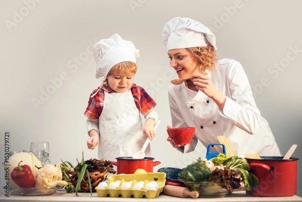 Fototapeta Mother and child chef cook cooking together. Kid chef cook helping mother to cooking. Mother and son cooking healthy food. Mother chef cook with apron and baby chef cook isolated in studio.