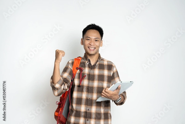 Fototapeta A cheerful young man in a plaid shirt, holding a tablet and backpack confidently, expressing enthusiasm and success. Perfect for themes of education, technology, achievement, and youthful energy.