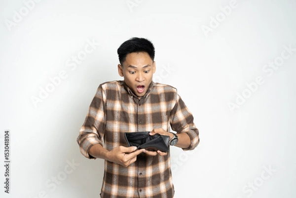 Fototapeta Surprised Asian Man Looking at an Empty Wallet on a White Background. Depicting financial concerns or unexpected expenses.