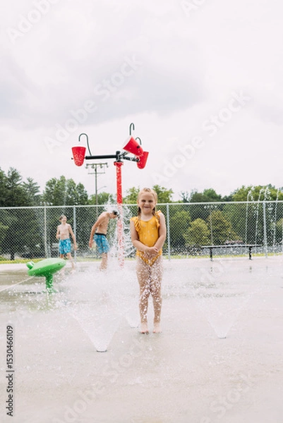 Obraz Smiling child standing in sprinklers while kids play in splash pad