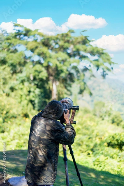 Obraz Man, photographer is wearing camouflage jacket taking photos with Dslr camera and telephoto lens in green forest at Khao Yai's 30th km Viewpoint in the Khao Yai National Park, Thailand.Vertical.