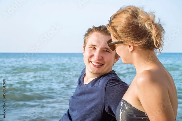Fototapeta A smiling young guy and a girl are talking on the beach against the background of the blue sea and blue sky on a sunny summer day