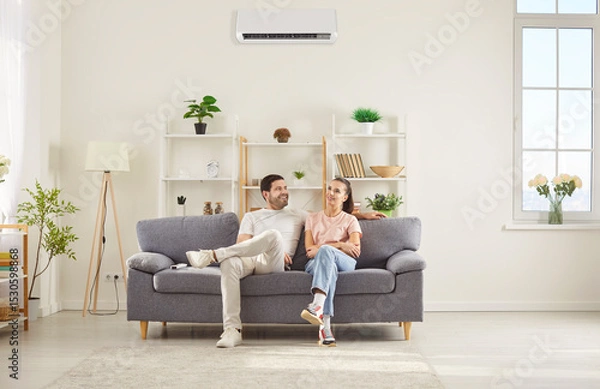 Fototapeta Happy couple sitting on comfortable gray sofa in living room, relaxing together under air conditioner. Smiling young man and woman resting on couch, talking and enjoying fresh cool air at home.