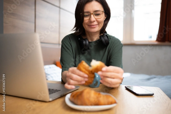 Fototapeta Pretty Woman Enjoys A Croissant For Breakfast While Checking Social Media On Laptop