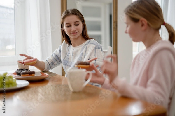 Fototapeta Two Smiling Sisters In Pajamas Have Breakfast With Donuts In A Kitchen