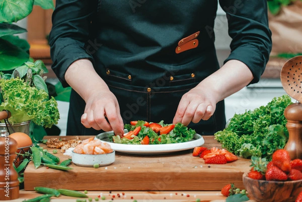 Fototapeta Chef carefully assembling vibrant salad with fresh greens, shrimps and strawberries on wooden countertop. Modern cozy kitchen filled with organic produce, creating inviting cooking atmosphere.