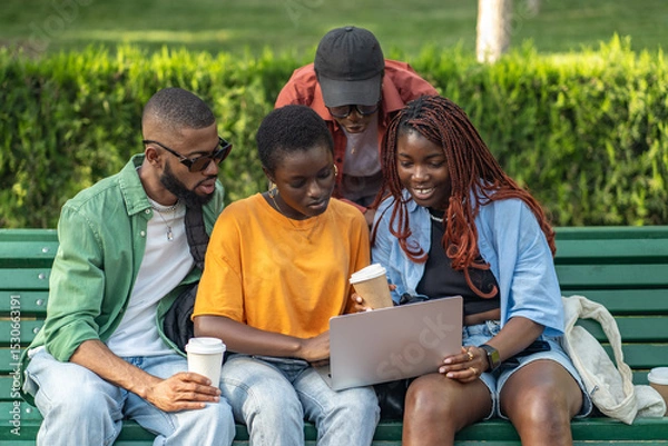 Fototapeta Group of African American friends enjoys coffee and work on laptop in campus park. Focus on homework, share jokes, watching movies and surfing internet, webinars for students, startup on freelance