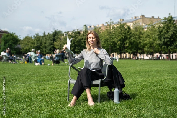 Fototapeta Joyful woman using makeshift paper fan to cool down, sunbathing on grass in hot summer weather. Businesswoman taking break, heat adaptation, solo relax, post work cooling, hydration with water bottle