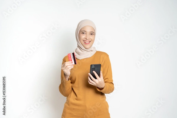 Fototapeta Confident woman holding a credit card and smartphone, expressing satisfaction and readiness for online activities such as shopping or banking.