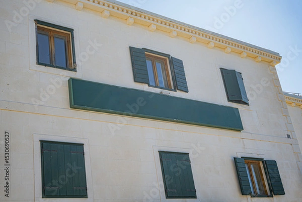 Obraz Facade of a traditional European stone building, featuring green wooden shutters on its multiple windows, all bathed in bright sunlight under a clear blue sky.