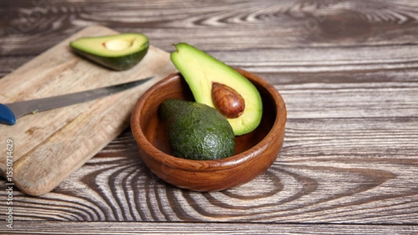 Fototapeta One halved avocado with seed and one whole in a wooden bowl, with a knife and avocado half on a cutting board over rustic wood surface