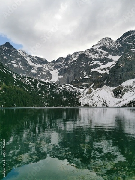 Fototapeta Towering snow-covered Tatra Mountains are reflected in the clear emerald waters of Morskie Oko lake beneath a moody sky near Zakopane, Poland