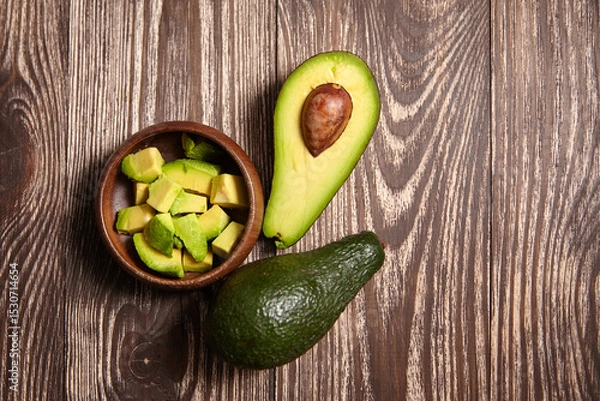 Fototapeta Fresh whole avocado, halved avocado with seed, and cubed pieces in a wooden bowl displayed on a dark rustic wooden background, perfect for food content, top view