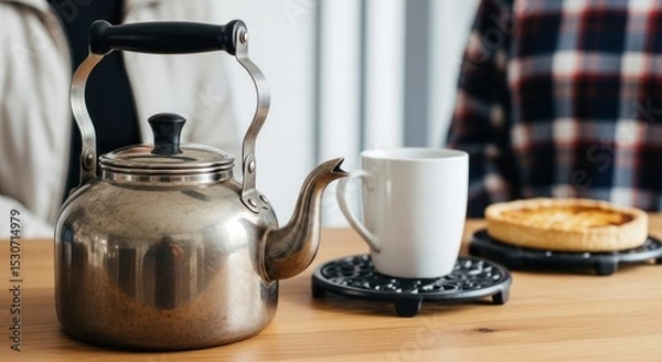 Obraz A stainless steel kettle and a white mug sitting on a wooden table with a pie in the background