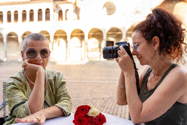 Obraz beautiful middle aged female couple having fun photographing themselves sitting at an outdoor table in a historic square