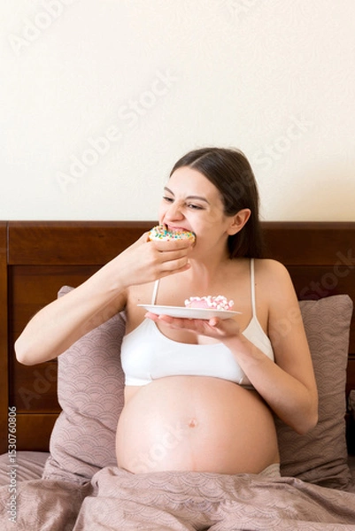Fototapeta Pregnant woman is eating many donuts relaxing in bed. Unhealthy dieting during pregnancy concept