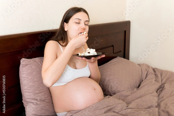 Fototapeta Hungry excited pregnant woman resting in bed is eating greedily a piece of cake and has a dirty mouth. Expecting mother can't stop eating
