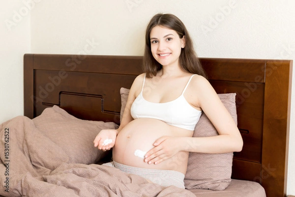 Fototapeta a pregnant girl sits at home on the bed and smears an anti-stretch mark cream on her stomach. Pregnancy, motherhood, preparation and expectation concept
