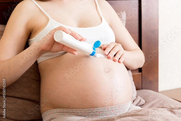 Fototapeta a pregnant girl sits at home on the bed and smears smile an anti-stretch mark cream on her stomach. Pregnancy, motherhood, preparation and expectation concept