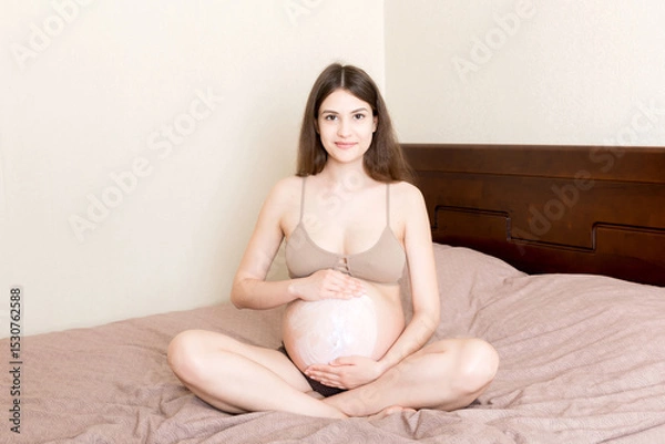 Fototapeta a pregnant girl sits at home on the bed and smears an anti-stretch mark cream on her stomach. Pregnancy, motherhood, preparation and expectation concept