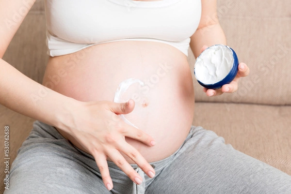 Fototapeta a pregnant girl sits at home on the bed and smears heart an anti-stretch mark cream on her stomach. Pregnancy, motherhood, preparation and expectation concept