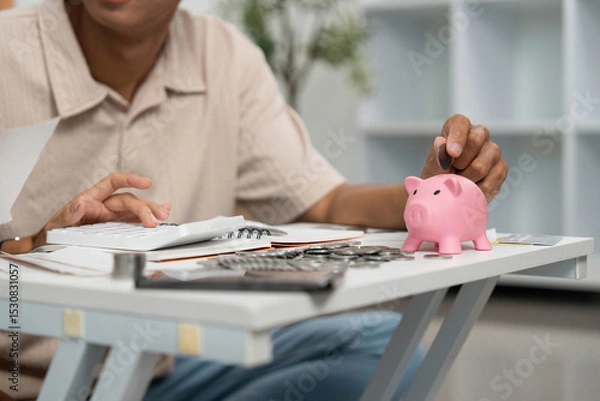 Obraz Financial Management and Savings. Young man depositing coins into a piggy bank at home.