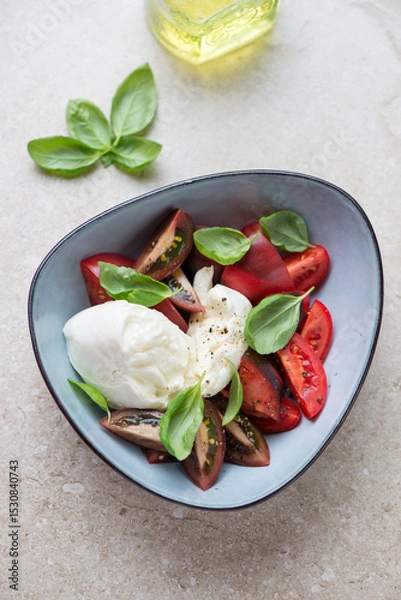 Fototapeta Italian caprese with buffalo mozzarella, tomatoes and fresh basil in a blue bowl, vertical shot on a beige stone background, high angle view
