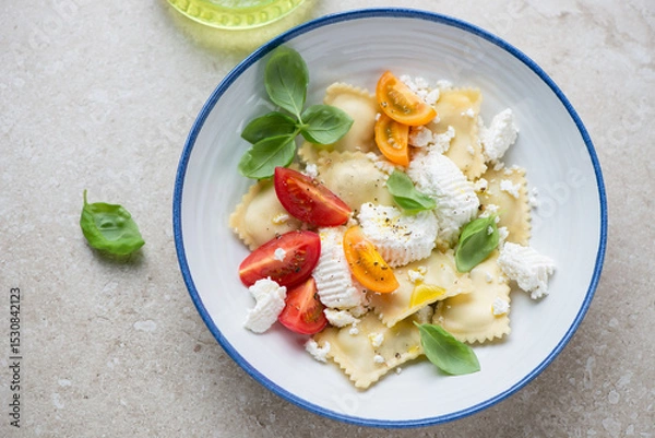 Fototapeta Italian ravioli served with ricotta, basil and tomatoes in a white and blue plate, horizontal shot on a beige stone background, elevated view