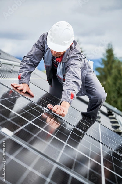 Fototapeta Man roofer mounting photovoltaic solar panels on roof of house. Engineer in helmet installing solar module system with help of hex key. Concept of alternative, renewable energy.