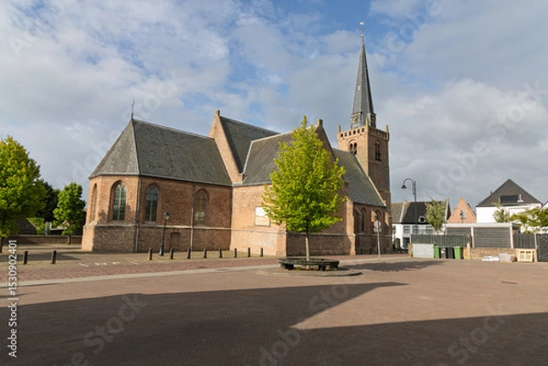 Obraz The Hervormde Kerk (Reformed Church) in Lexmond, Netherlands, stands to historical Dutch architecture. Its Gothic style, red brick, and tall spire dominate the village skyline under a serene sky.
