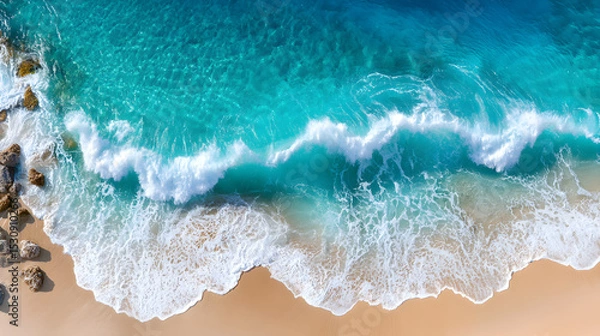 Obraz Aerial view of turquoise ocean waves gently crashing onto a sandy beach, with rocky formations enhancing the natural coastal landscape. Selective focus