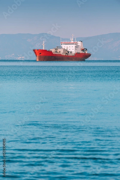 Fototapeta Red cargo ship sailing on calm blue sea with mountains in background, maritime transport concept