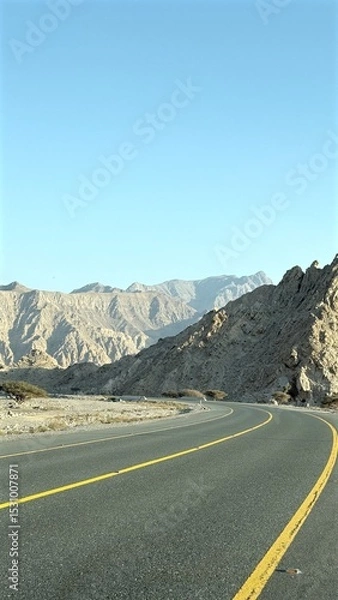Fototapeta Road curves beneath towering cliffs as shadows ripple across rugged terrain, cliffs rise sharply from ancient layers, forming deep rhythm carved into raw geological landscape.