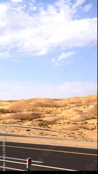 Fototapeta Curving asphalt road threads across wind-brushed dunes under painted sky, bright yellow line holding its course through soft sand that creeps onto shoulder, showing tension between nature and