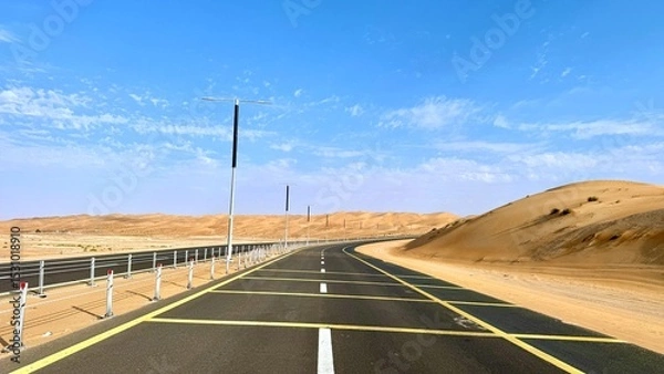 Fototapeta Road slices through golden sand as poles line both sides, road reflects heat under vibrant sky while sand dunes rise gently, creating harmony between built line and desert stillness.