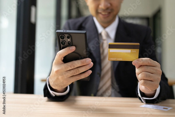 Fototapeta A man is using a laptop and a cell phone to pay for something. He is holding a credit card and a debit card
