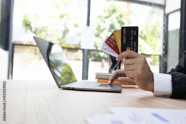 Fototapeta A man is using a laptop and a cell phone to pay for something. He is holding a credit card and a debit card
