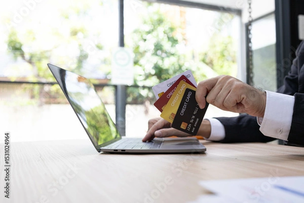 Fototapeta A man is using a laptop and a cell phone to pay for something. He is holding a credit card and a debit card
